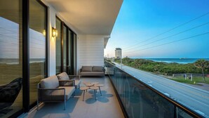 A balcony with a sofa, two small tables, and a view of the ocean and a road. - Ocean's Edge | Lavish NEW Condo w Beach Views, Cozy Decor & Community Amenities (Melbourne Beach)