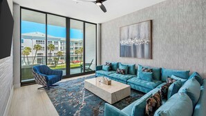 A modern living room with a large sofa set, a coffee table, and a ceiling fan. There is a large window offering a view of buildings and palm trees. - Ocean's Edge | Lavish NEW Condo w Beach Views, Cozy Decor & Community Amenities (Melbourne Beach)