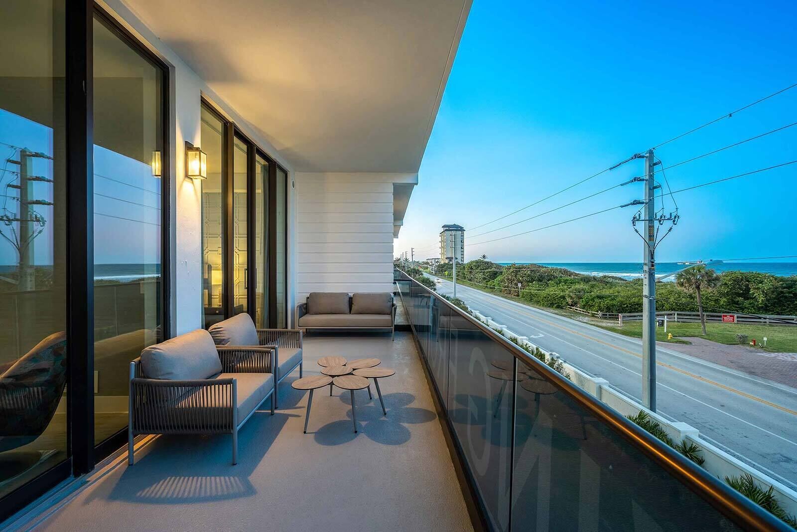 A balcony with a sofa, two small tables, and a view of the ocean and road.