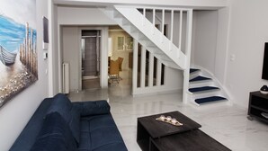 A modern living room with a dark blue sofa, a coffee table with decorative items, a white staircase, and a view into the dining area. - Garitsa Gem Suite (Corfu)