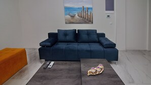 A modern living room with a dark blue sofa, an orange ottoman, and a coffee table with decorative items. - Garitsa Gem Suite (Corfu)