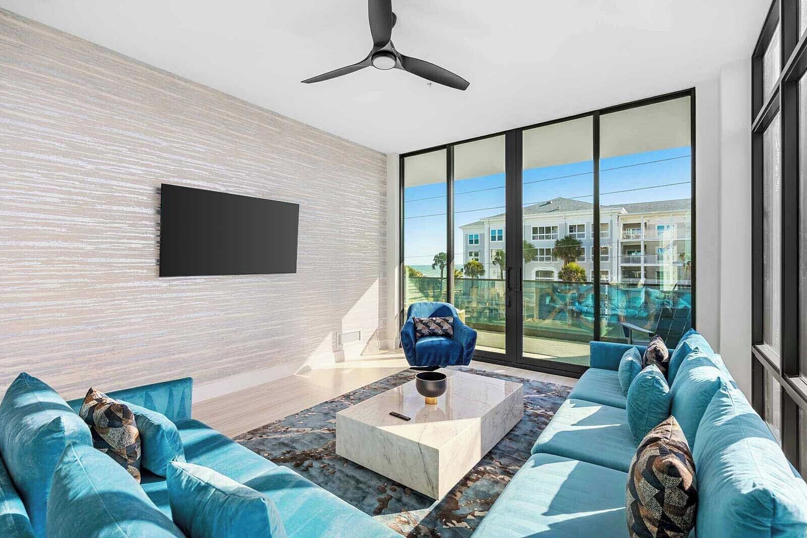 A modern living room with a large window, a ceiling fan, and a flat-screen TV mounted on the wall.