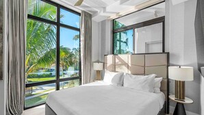 A modern hotel room with a large bed, two bedside tables with lamps, a ceiling fan, and large windows offering a view of palm trees and a pool.