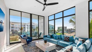 A modern living room with a large sofa, a coffee table, and a chair. The room has large windows offering a view of buildings and palm trees.