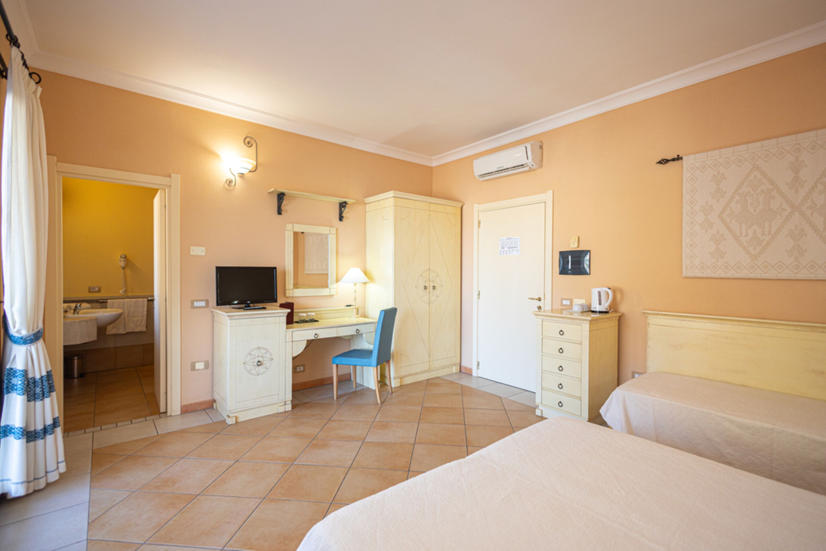 A hotel room with a bed, a desk with a chair, a television, a wardrobe, and an air conditioning unit.