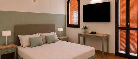 A bedroom with a bed, bedside tables, a desk, a flat-screen TV, and a window with blinds.