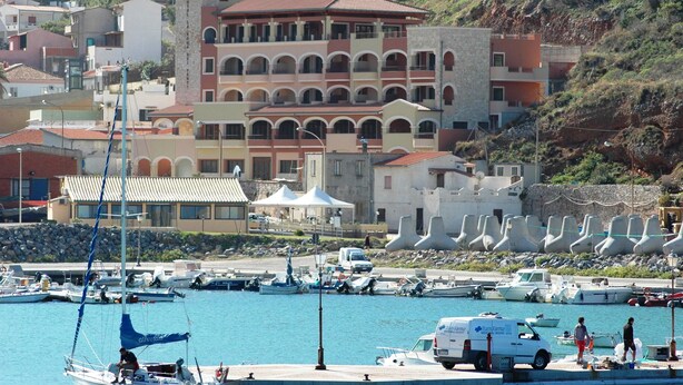 Una marina costera con barcos, una camioneta blanca y un edificio con varios balcones.