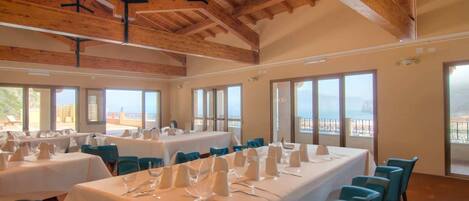 Un comedor con mesas dispuestas para una comida, ventanas amplias con vistas y vigas de madera en el techo.
