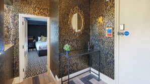 A hallway with patterned wallpaper, a mirror, a door with a sign, and a bed in the adjacent room.