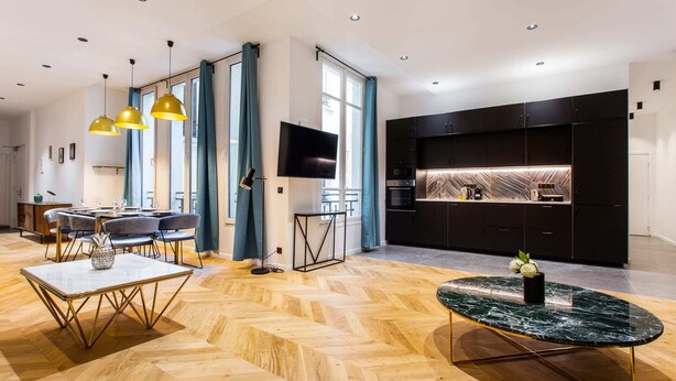 A modern living room with a herringbone wood floor, a marble coffee table, and a dining area with a white table and gold chairs. - Résidence Paris Boetie (Paris)
