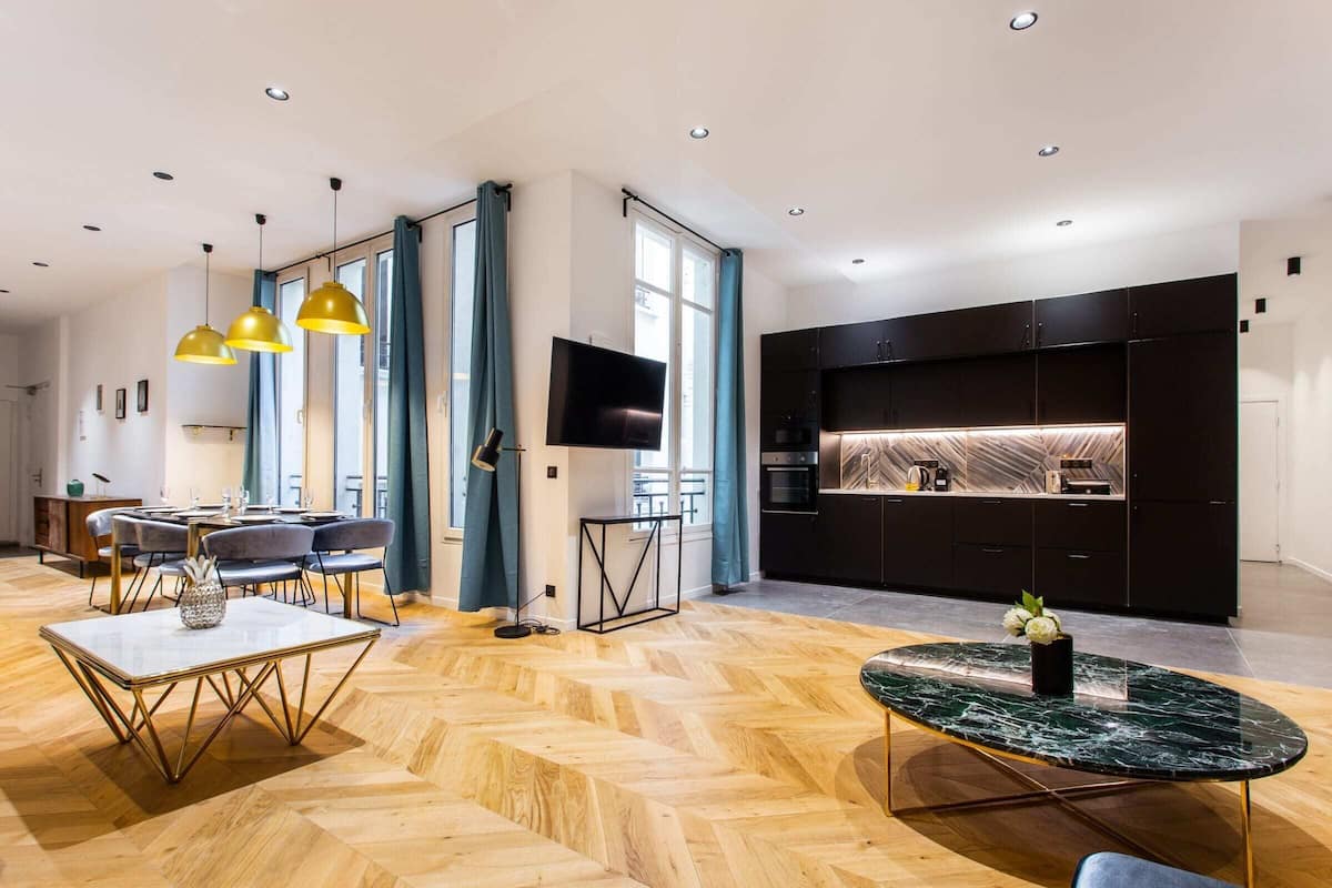 Uma sala de estar moderna com piso de madeira em padrão herringbone, uma mesa de centro de mármore e uma área de jantar com mesa branca e cadeiras douradas.