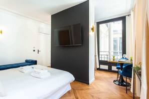 Habitación de hotel con cama, televisión montada en la pared, zona de comedor con mesa y sillas, y ventana con vistas.