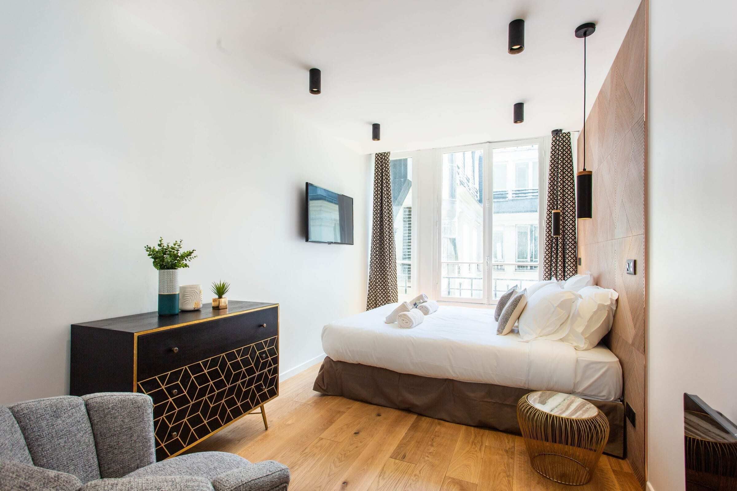 A modern bedroom with a large bed, a dresser, a chair, and a small round ottoman.