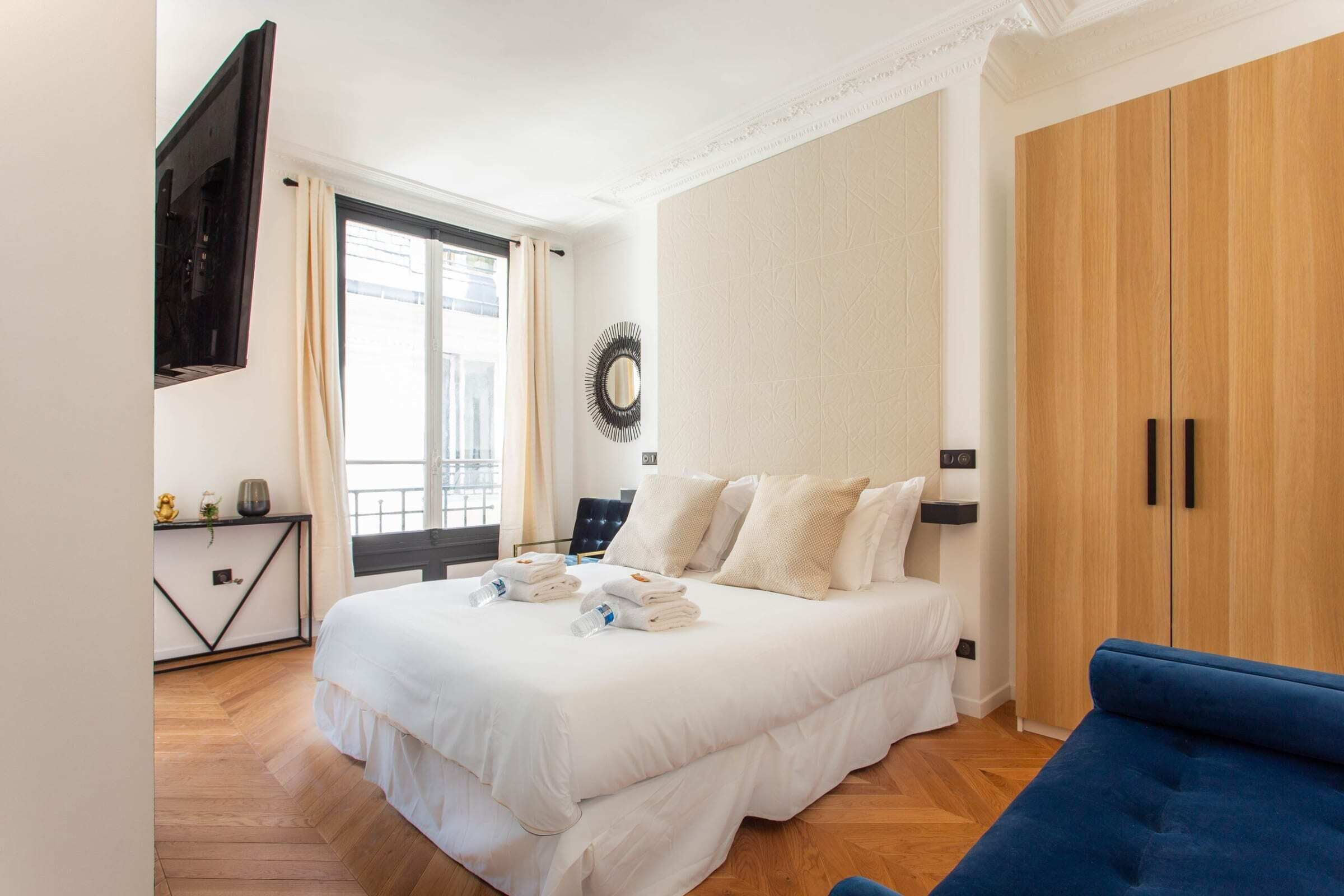 A hotel room with a bed, a blue sofa, a wardrobe, a TV, a window with curtains, and a wall-mounted mirror.