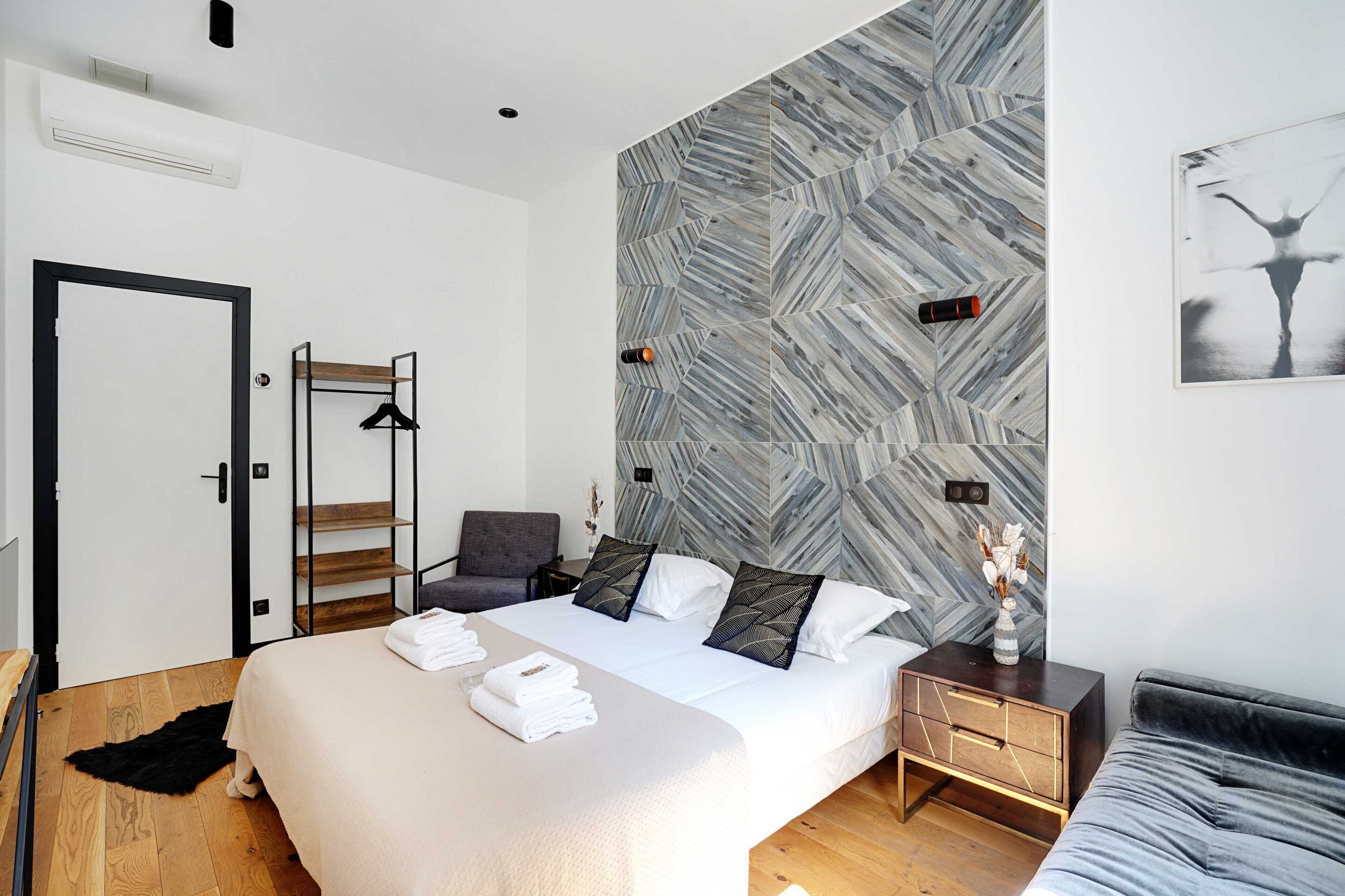 A modern bedroom with a large bed, a wooden floor, a grey sofa, a wooden nightstand, and a wall with a geometric pattern.