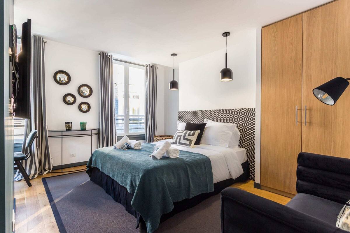 Quarto de hotel moderno com cama bem arrumada, uma secretária com cadeira, guarda-roupa e uma janela grande com cortinas.