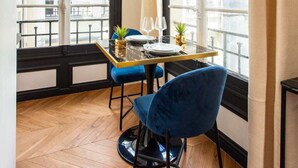 A dining area with a table set for two, blue chairs, and a view of the city through large windows. - Résidence Paris Boetie (Paris)