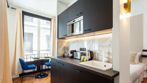 A compact kitchen with a built-in oven, a coffee maker, and a sink. There is a dining area with blue chairs and a table. - Résidence Paris Boetie (Paris)