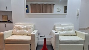 Uma sala de estar moderna com dois sofás de couro branco, uma mesa pequena vermelha e uma janela com cortinas.