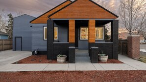 Une maison moderne à deux étages, dotée d’un porche en bois à l’avant, de détails en briques noires et d’un petit jardin agrémenté de paillis et de plantes en pot.