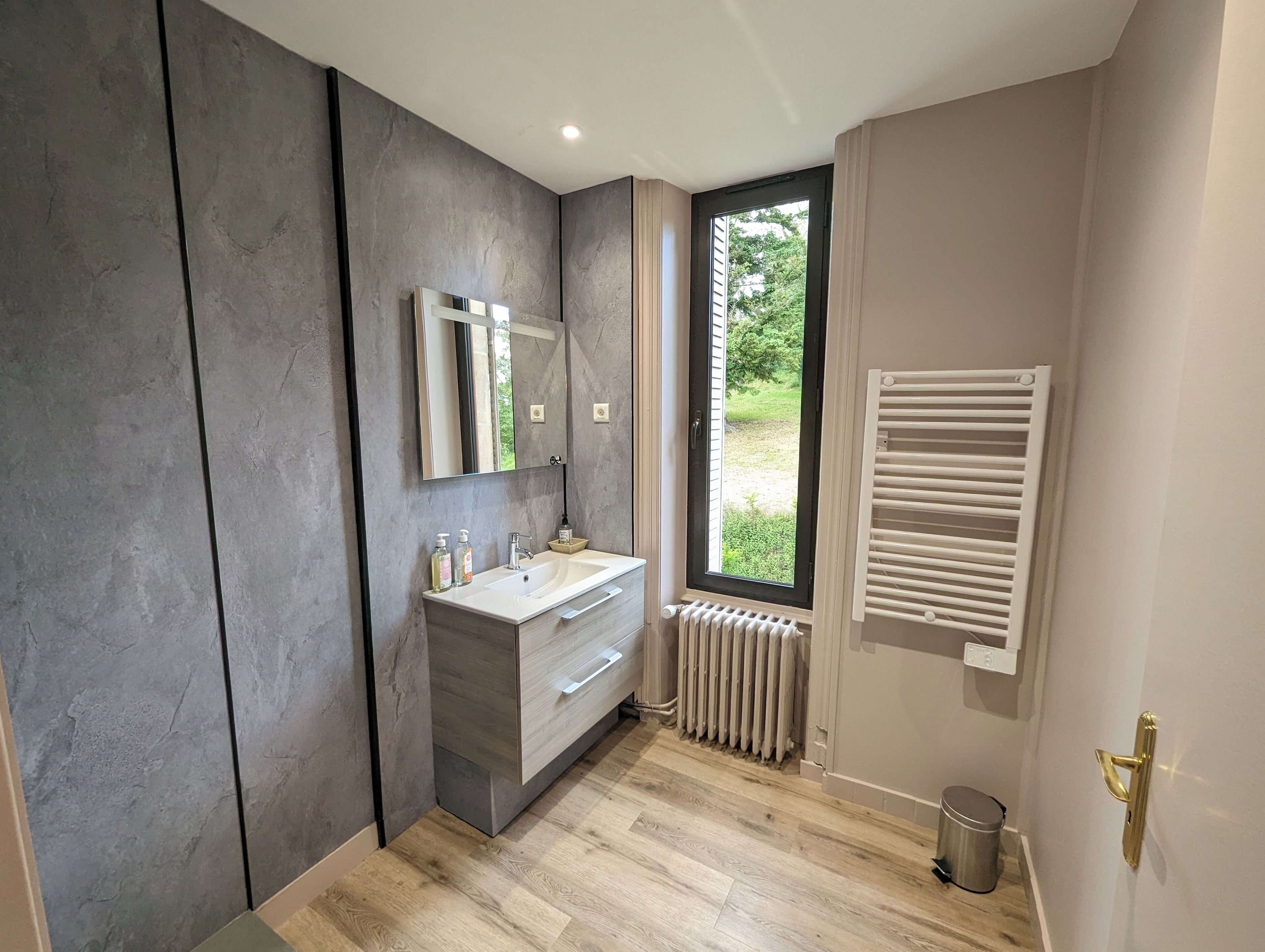 Ein modernes Badezimmer mit einem großen Spiegel, einem weißen Waschbecken und einem Fenster mit Blick auf Grünflächen.