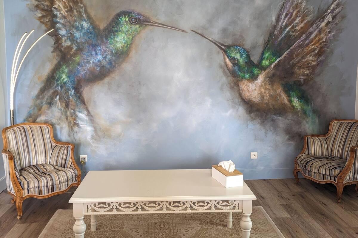 A living room with a large abstract painting of hummingbirds, two striped armchairs, a white table, and a blue sofa.