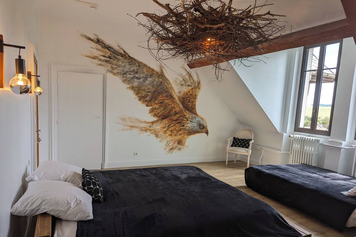 A bedroom with a sloped ceiling, a mural of an eagle, two beds, a chair, and a window.