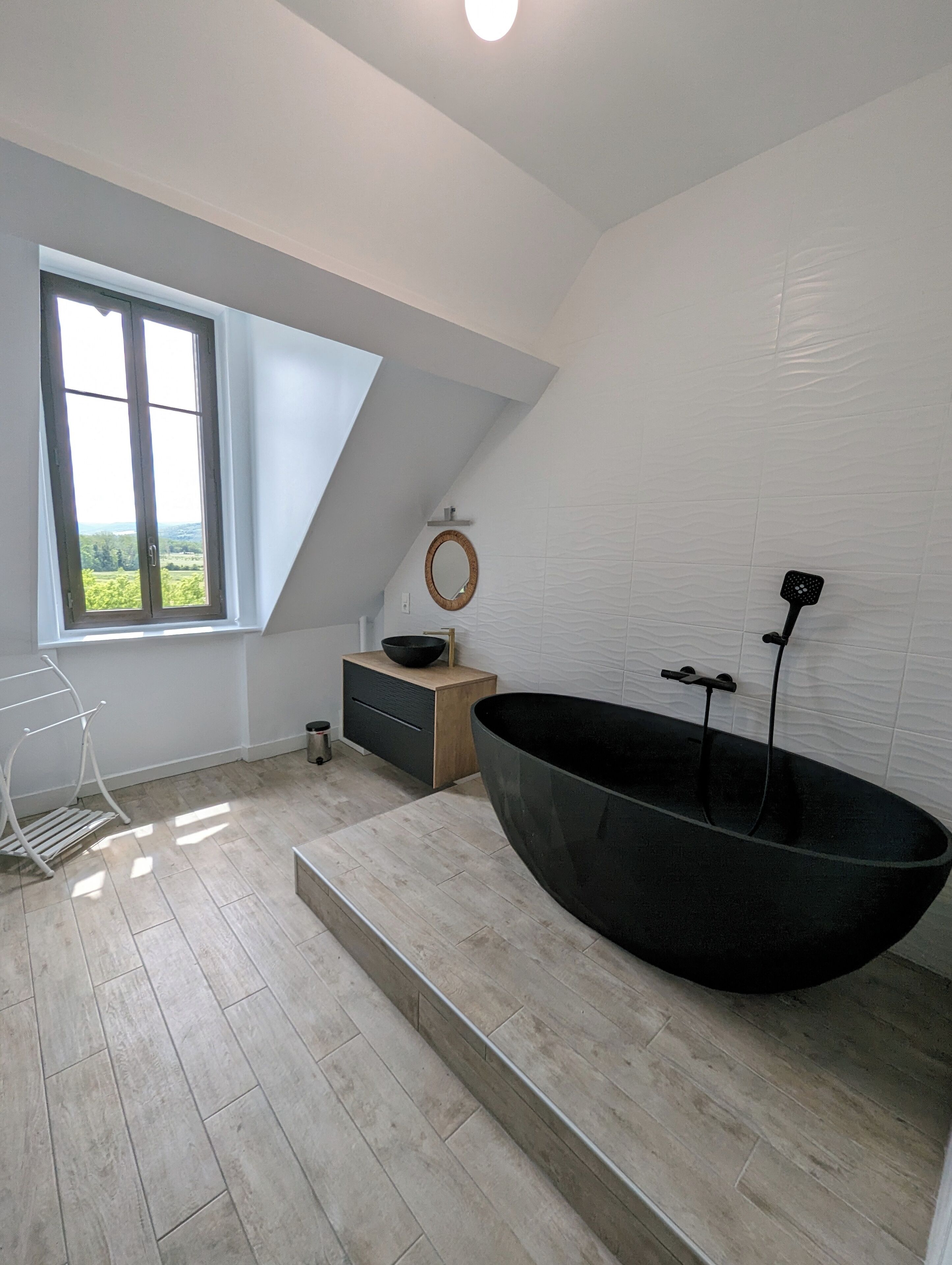 Une salle de bain moderne avec une grande baignoire noire, un meuble-lavabo en bois, un petit lavabo et une fenêtre donnant sur un paysage verdoyant.