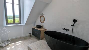 Une salle de bain moderne avec une grande baignoire noire, un meuble-lavabo en bois, un petit lavabo et une fenêtre donnant sur un paysage verdoyant.