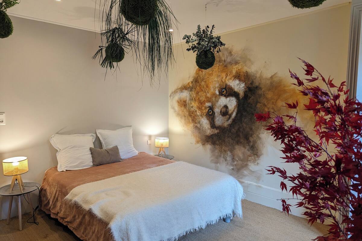 A bedroom with a large bed, two bedside lamps, a wall mural, and hanging plants.
