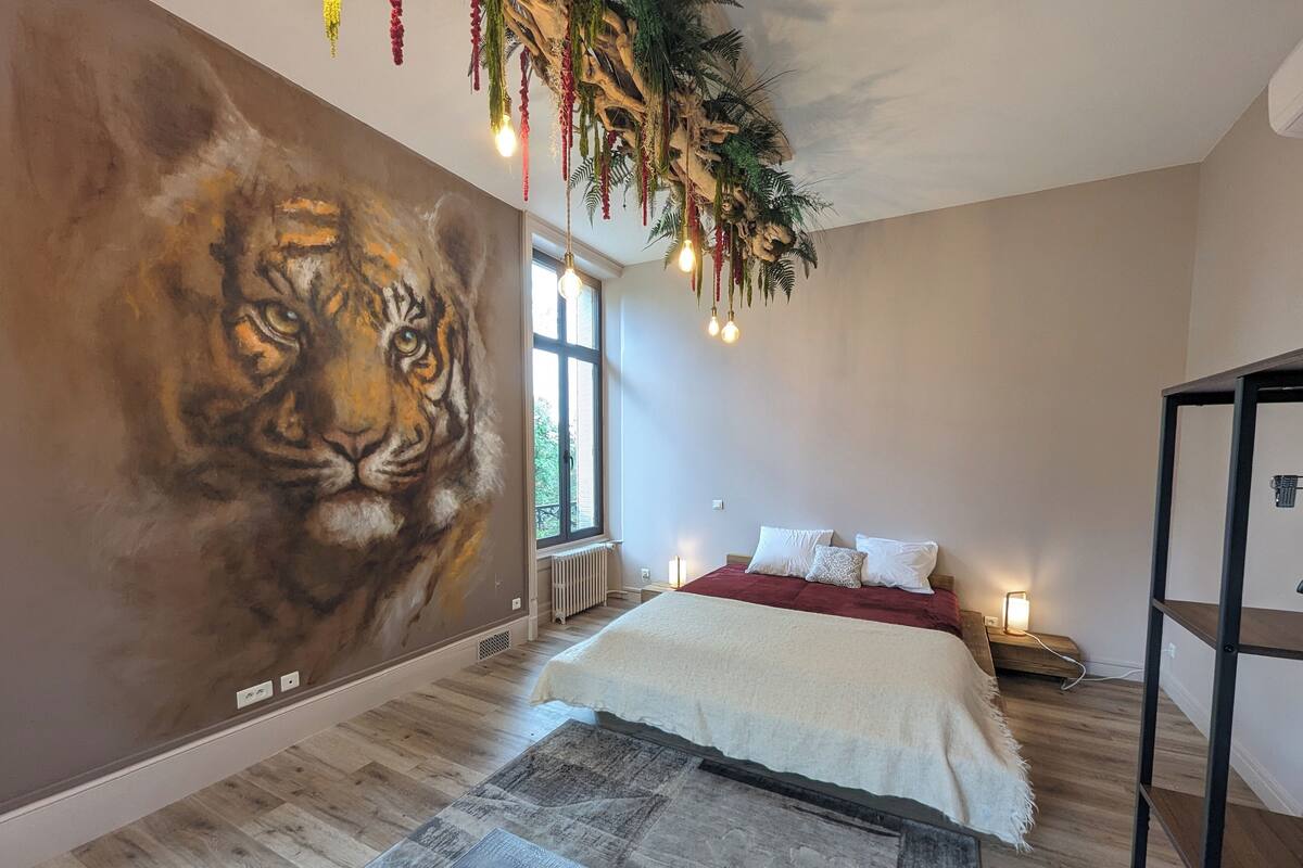 A bedroom with a large wall mural of a tiger, a bed with white bedding, a wooden floor, and a hanging plant with lights.
