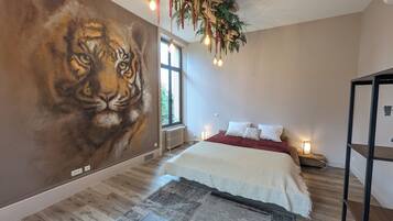 A bedroom with a large wall mural of a tiger, a bed with white bedding, a wooden floor, and a hanging plant with lights.