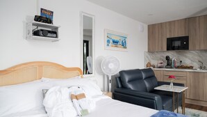 Quarto de hotel com cama, poltrona, pequena mesa com um vaso, micro-ondas e cozinha.