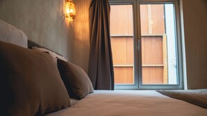 Una cama bien tendida con almohadas, una lámpara fijada a la pared y vistas a un balcón de madera a través de una ventana.