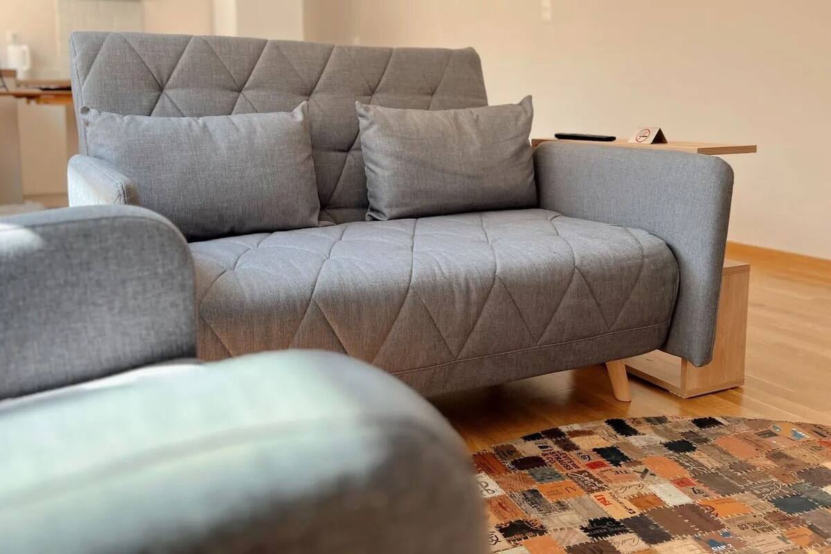 A grey sofa with a tufted backrest and armrests, positioned on a patterned rug in a room with wooden flooring.