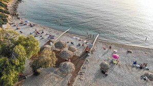 Ovenfra ses en strand med folk, der solbader og svømmer, hytter med stråtag og en mole, der stikker ud i vandet.