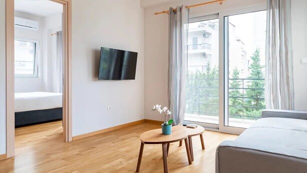 Et moderne hotelværelse med trægulv, seng, tv ophængt på væggen, et lille bord med en vase blomster og en balkon med gardiner.