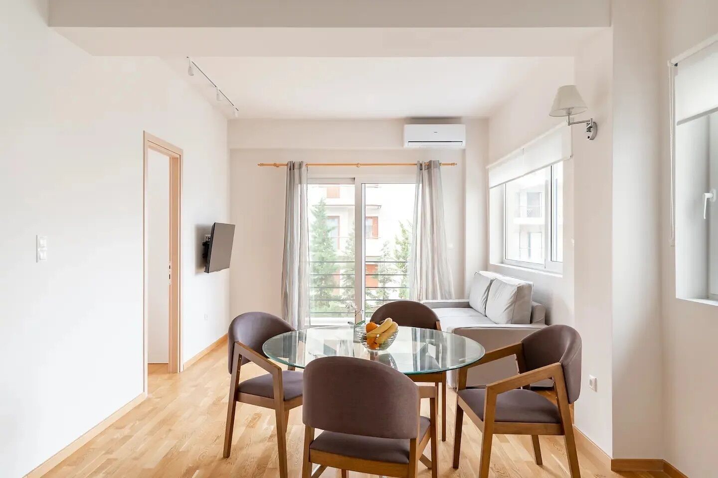 A modern living room with a glass dining table, four chairs, a sofa, and a TV mounted on the wall.