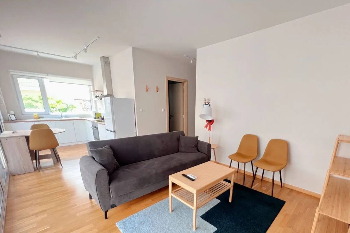 A modern living room with a grey sofa, wooden coffee table, and two armchairs. A kitchen with white cabinets and a dining area with a small table and chairs is visible in the background.