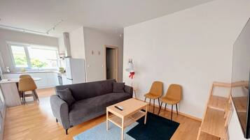 Una sala de estar moderna con un sofá gris, una mesa de centro de madera y dos sillones. Se ve una cocina con gabinetes blancos y una zona de comedor con una mesa pequeña y sillas al fondo.