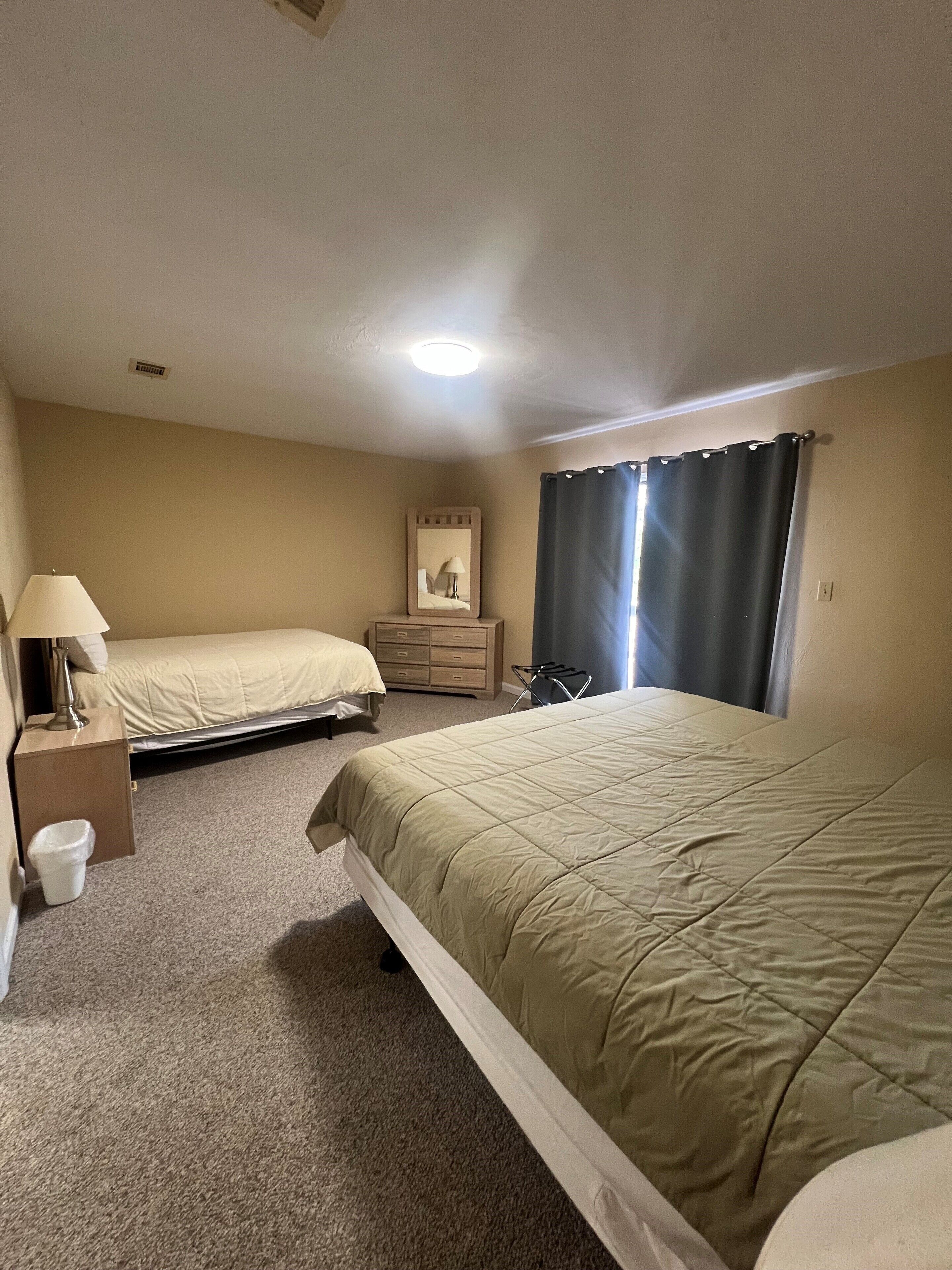 A hotel room with two beds, a dresser, a mirror, a lamp, and a window with curtains.