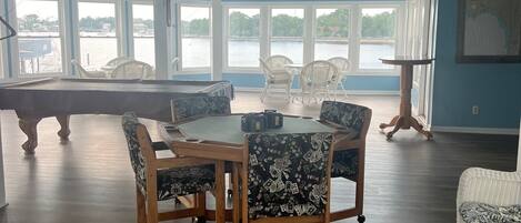 A spacious room with a pool table, a round dining table with chairs, and large windows offering a view of a body of water.