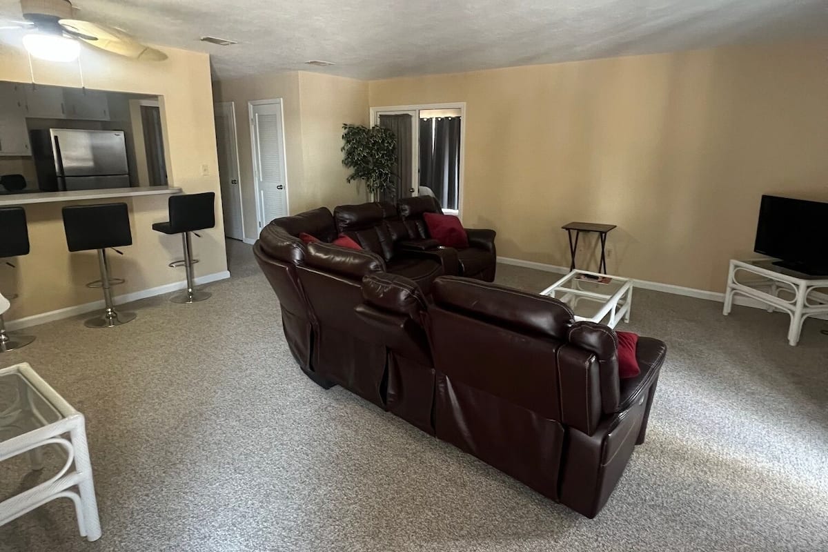 Ein Wohnzimmer mit braunem Ledersofa, einem kleinen weißen Tisch, einer TV-Konsole und einer Küchenzeile mit Kühlschrank.