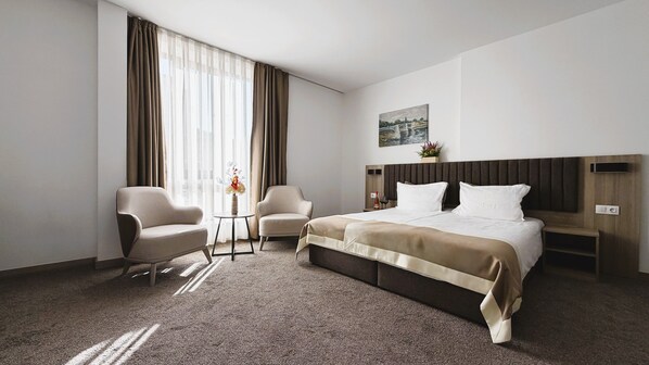 Et hotelværelse med en seng, to lænestole, et lille bord med en vase og et billede på væggen.