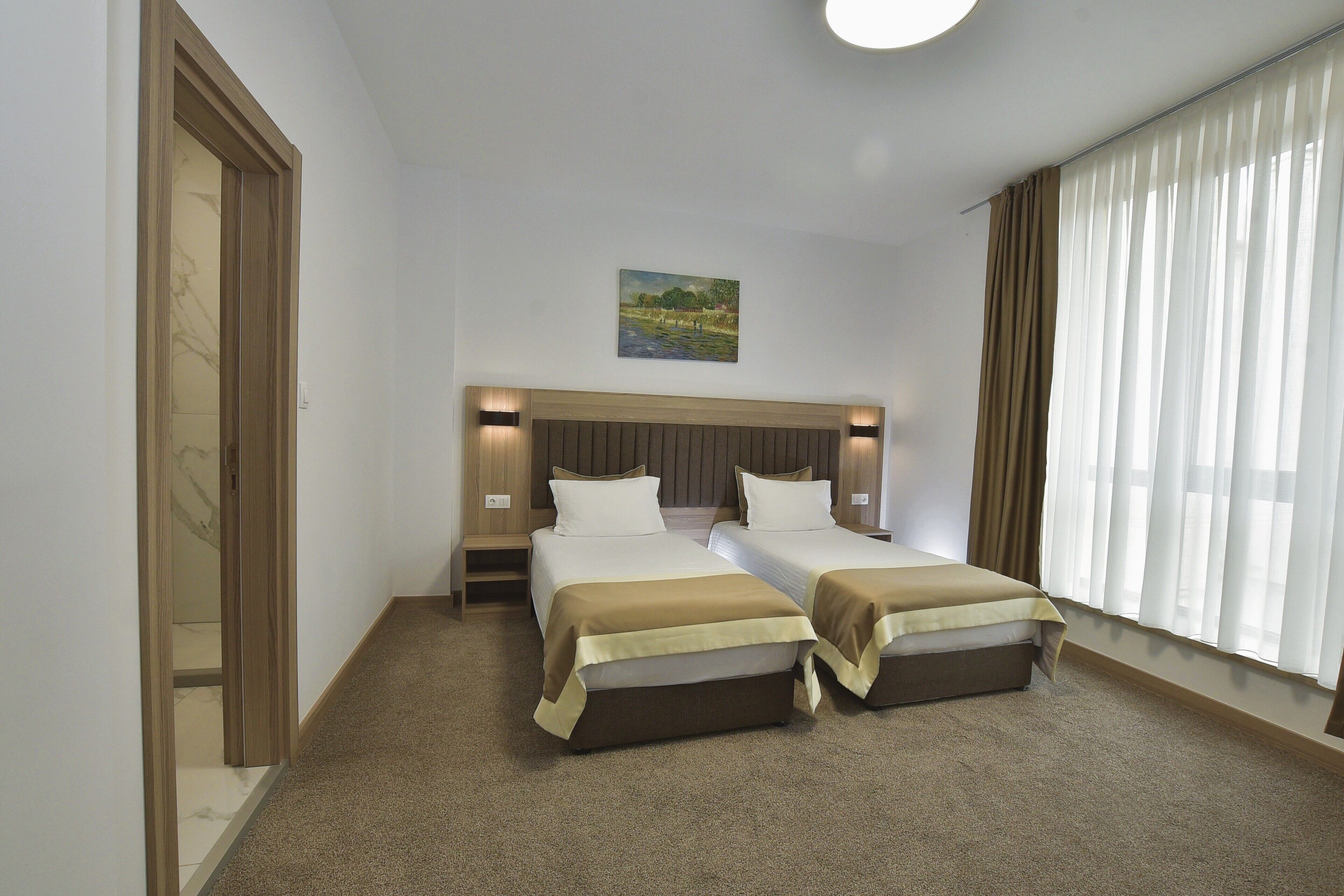A hotel room with two beds, a painting on the wall, and a large window with curtains.