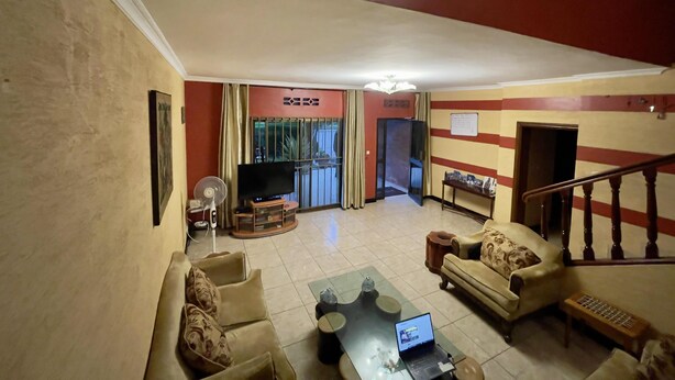 A living room with a sofa, a coffee table, a TV, and a laptop. There is a staircase with a railing leading to another level. - Captivating 3-bed House in Kigali , Rwanda (Kigali)