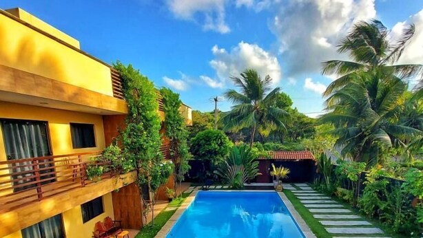 A swimming pool with a wooden deck and palm trees, surrounded by a yellow building with balconies. - Harmonia Flats (Tibau do Sul)