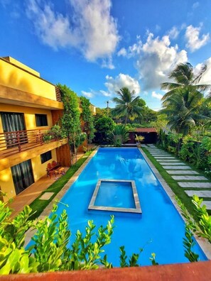 A swimming pool with a wooden deck and palm trees, surrounded by a yellow building with balconies. - Harmonia Flats (Tibau do Sul)