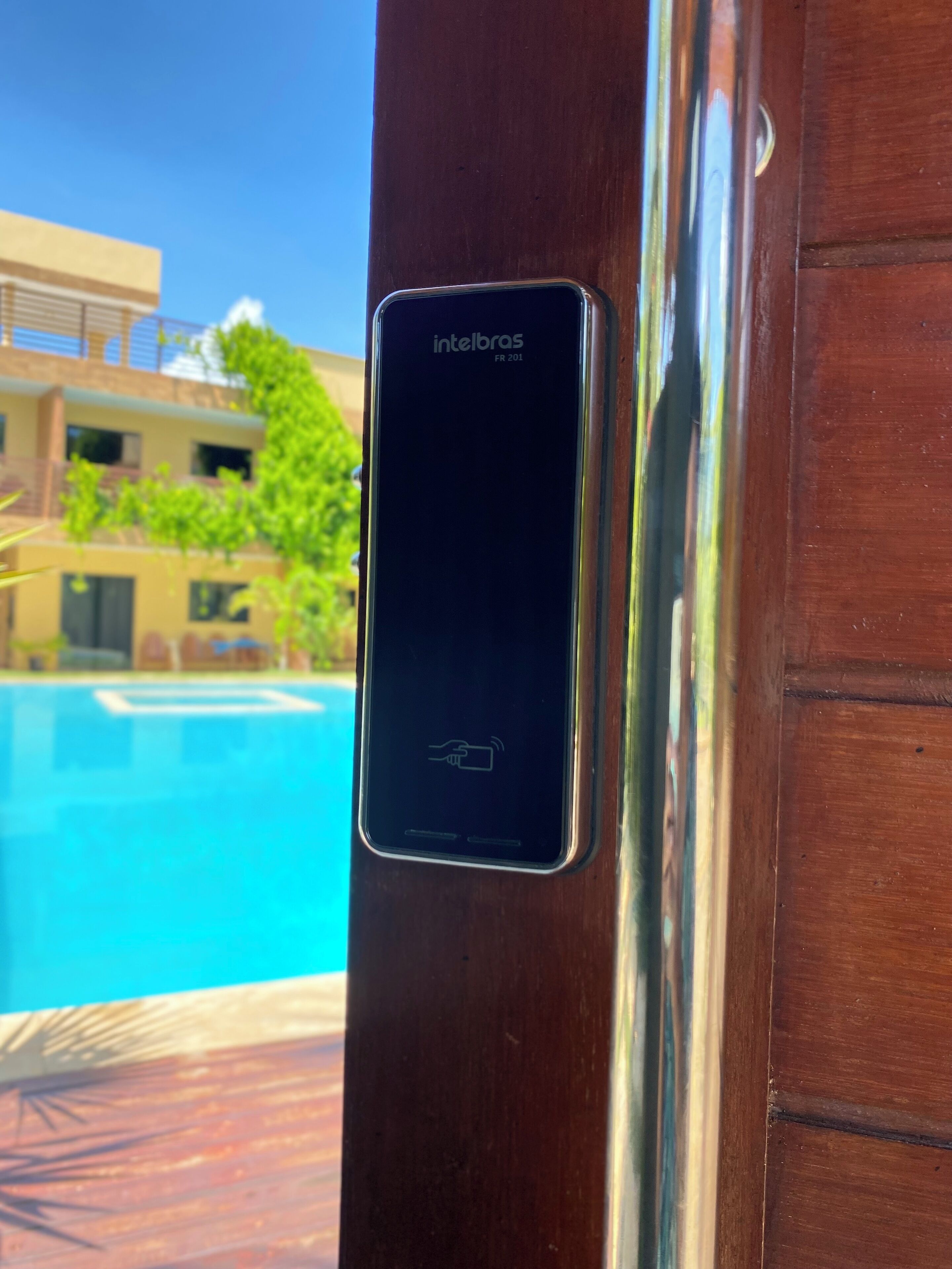 Un teléfono inteligente con la marca 'intelbras' montado en una puerta de madera, con una piscina y edificios al fondo.