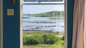 A room with a large window overlooking a coastal landscape, a cushioned bench, and a power outlet on the wall. - Canna Campsite (Isle of Canna)